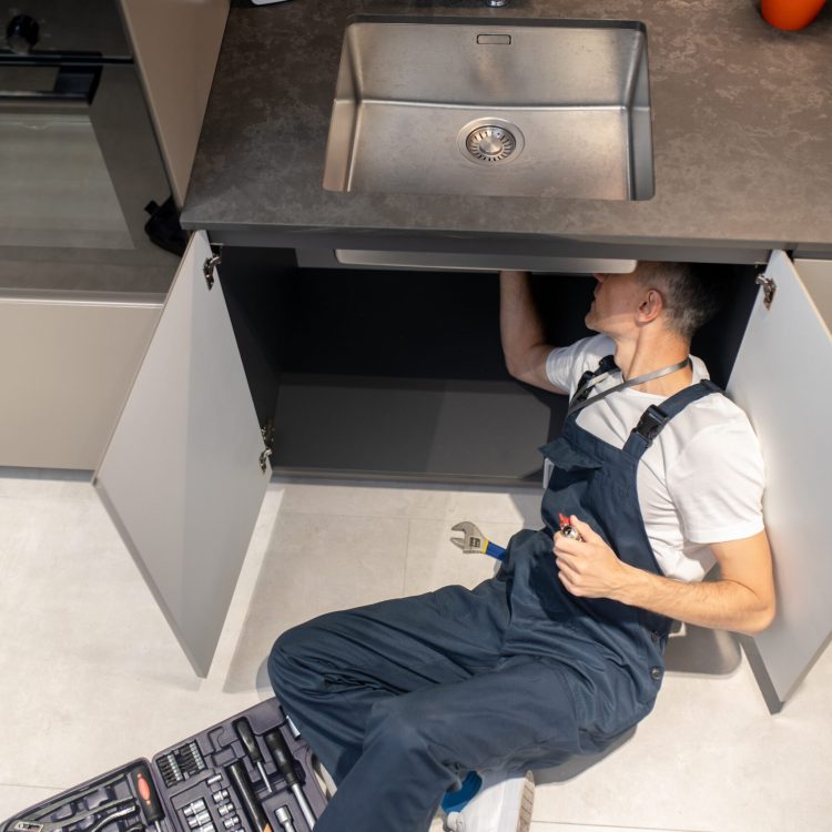 Man lying down fixing breakdown of sink Solution. Top view of man plumber in overalls holding ball valve in hand lying on floor near open set of tools in kitchen fixing breakdown of sink